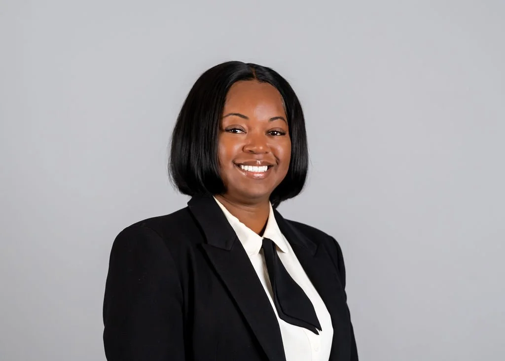 Professional woman with shoulder-length black hair wearing a black blazer, white shirt, and black tie, smiling in front of a plain gray background.