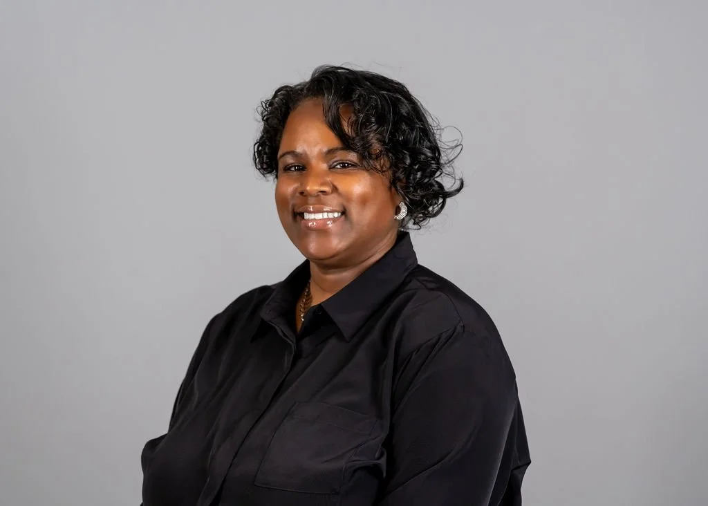Mid-age woman with short, curly black hair, smiling, wearing a black blouse, pearl earrings, and a necklace, against a light gray background.