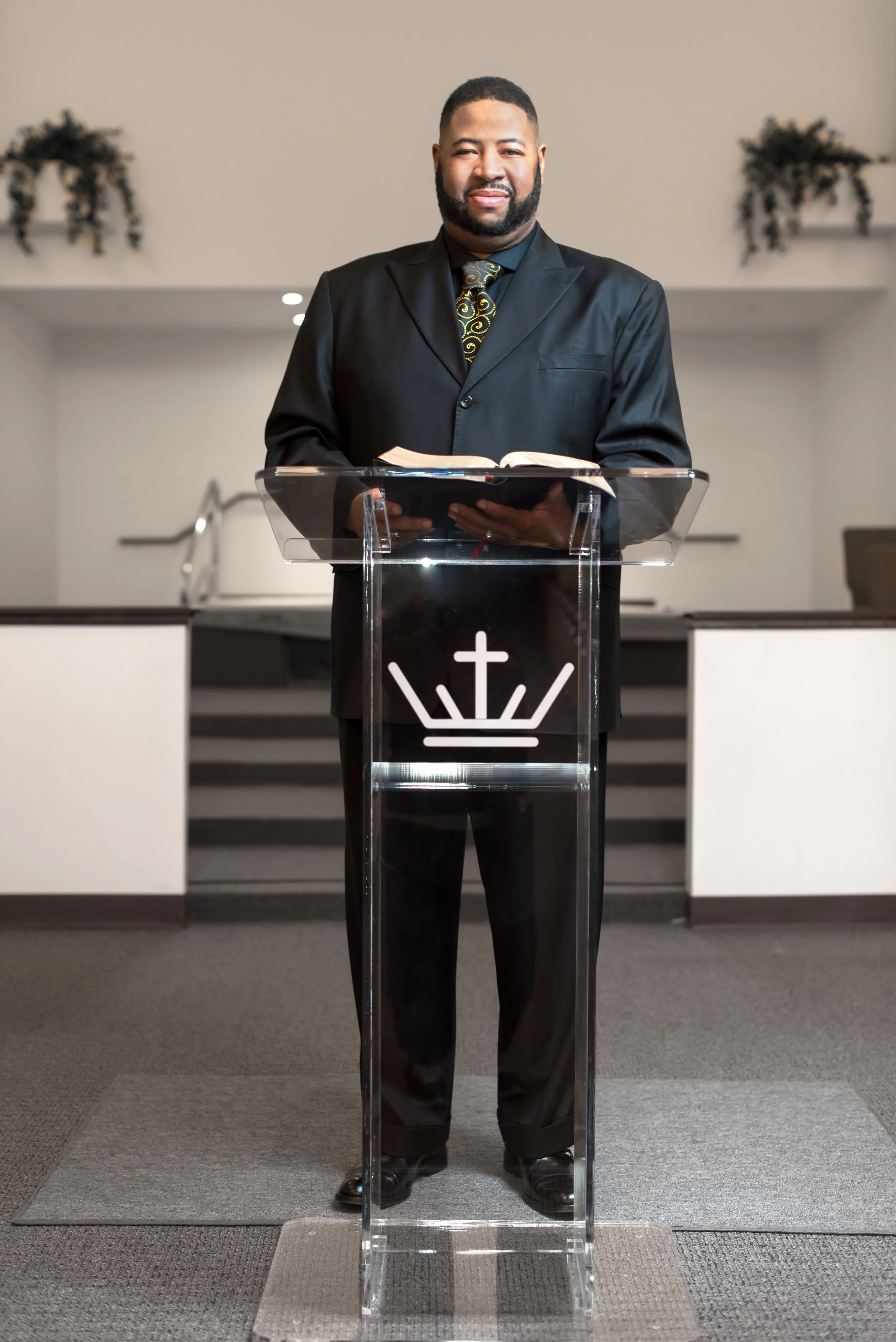A man in a black suit and patterned black and gold tie standing behind a clear pulpit with a cross and crown symbol in a church setting, with steps and decorative plants in the background.