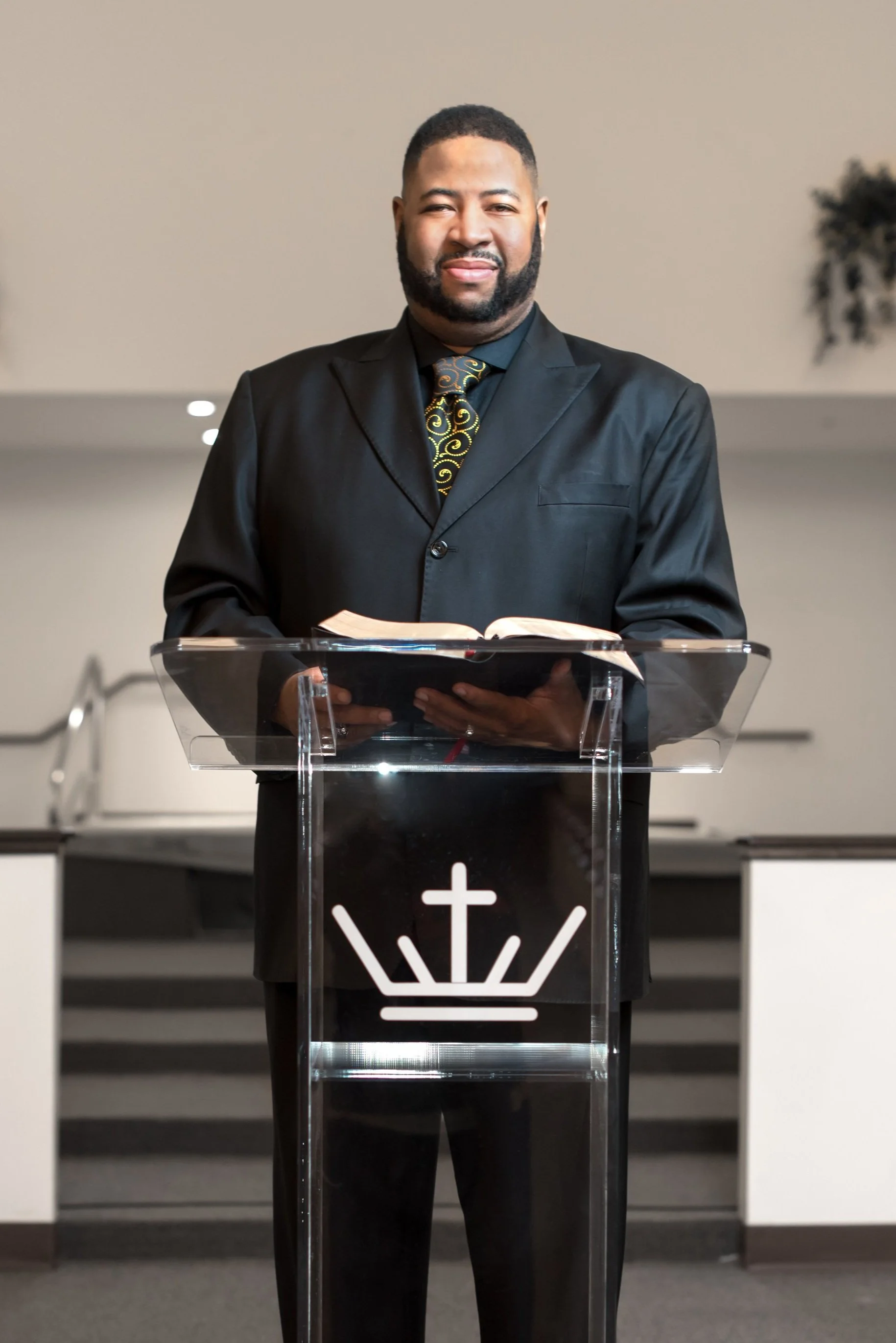 A man standing behind a clear lectern with a cross symbol, holding an open Bible, dressed in a black suit and tie, in a church or religious setting.