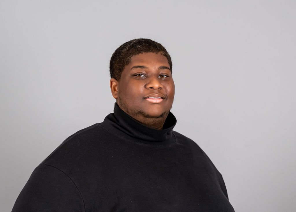 Portrait of a young man with short hair wearing a black turtleneck, standing against a plain light gray background.