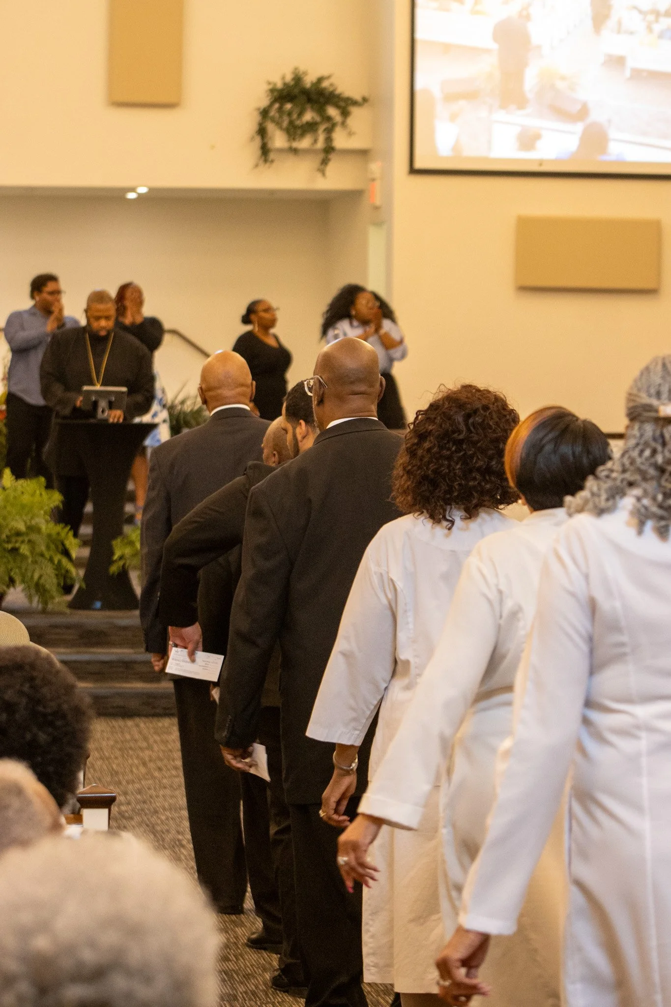 People standing in line during a formal event, with some holding papers, in a room with a large screen and people presenting at a podium.