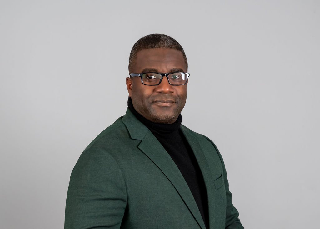 A man with short hair, glasses, wearing a dark green blazer and black turtleneck, looking at the camera with a confident expression, against a plain light gray background.