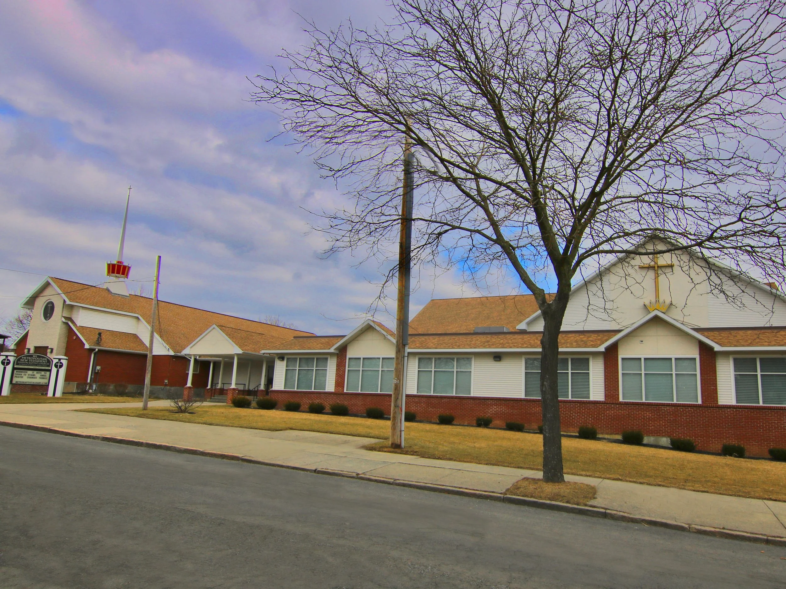A church building with a large cross on the front, surrounded by a lawn with small bushes, a sidewalk, and a leafless tree in front. The sky is partly cloudy.