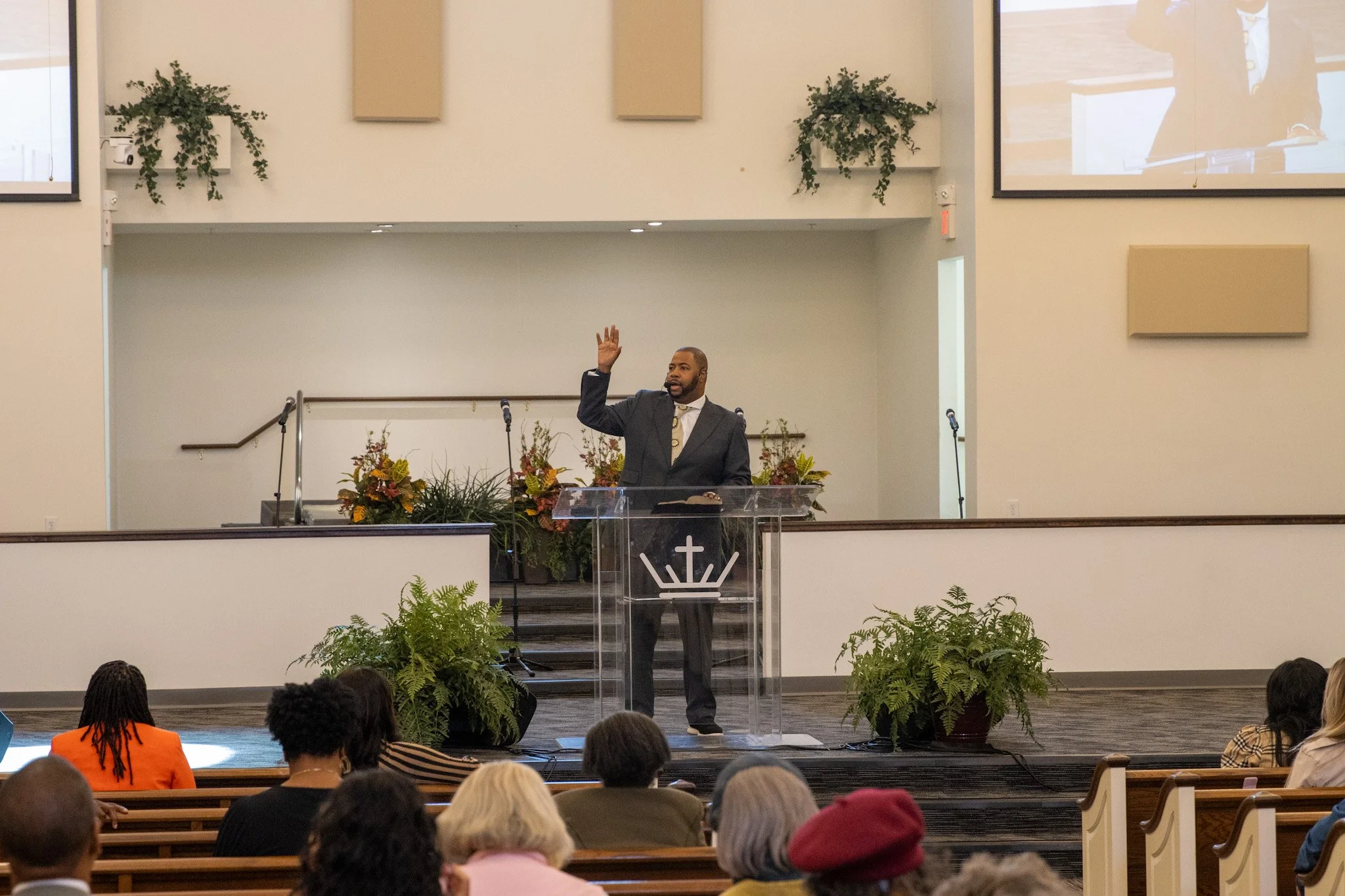 A man in a suit standing behind a clear pulpit, raising his right hand, in a church with an audience seated in pews. The church has large screens, plants, and flowers near the pulpit.