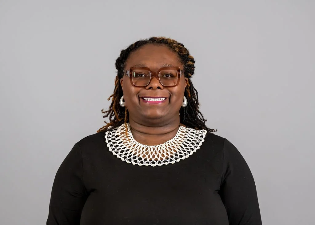 A woman wearing glasses, earrings, a necklace, and a black top, smiling and standing against a plain gray background.