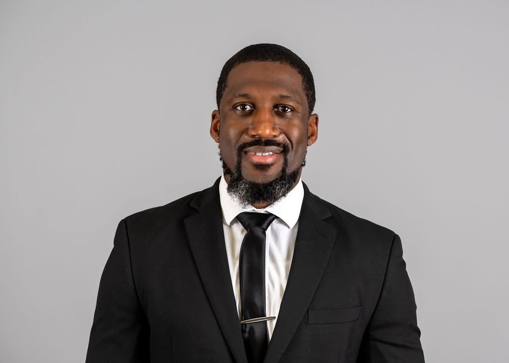 A man in a black suit with a white shirt and black tie, smiling against a plain gray background.