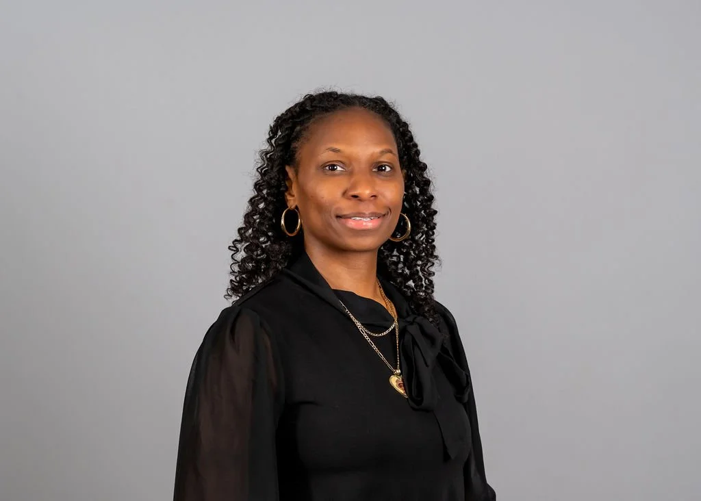 A woman with curly black hair, wearing a black blouse, gold hoop earrings, and gold necklaces, standing against a plain gray background.