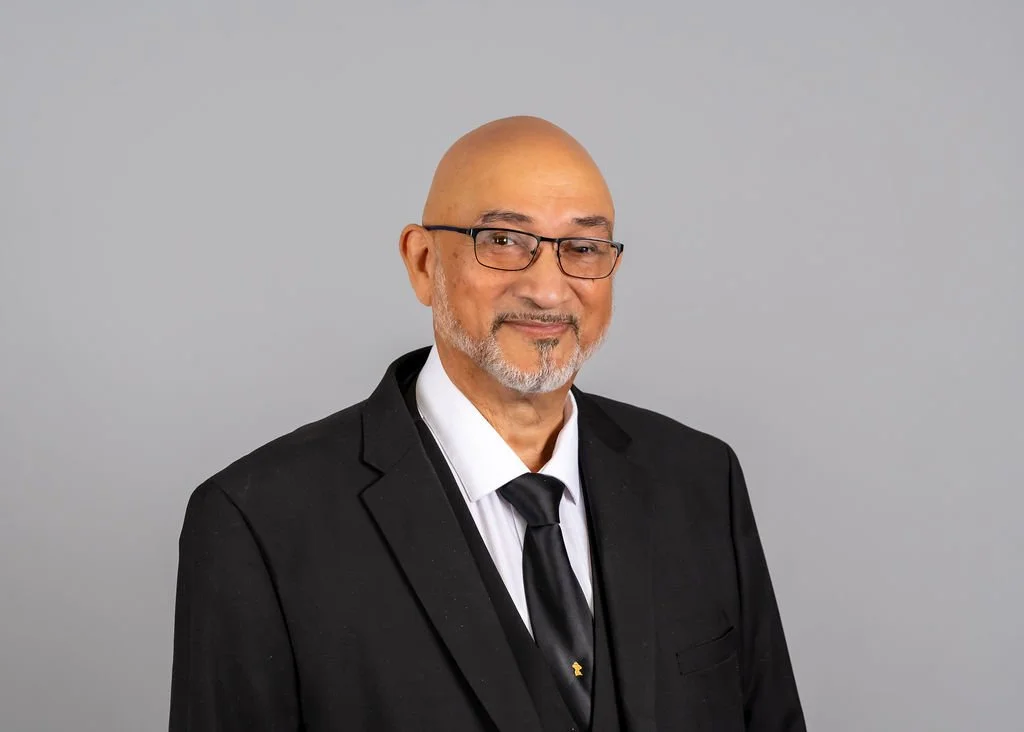 Headshot of an older man with glasses, a bald head, gray goatee, wearing a black suit, white shirt, and black tie, smiling slightly against a gray background.