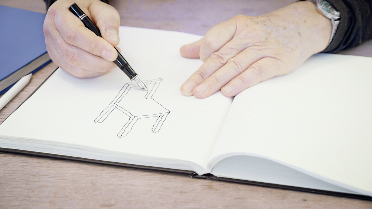 Person drawing a simple outline of a chair in a notebook with a black pen, sitting at a wooden table.