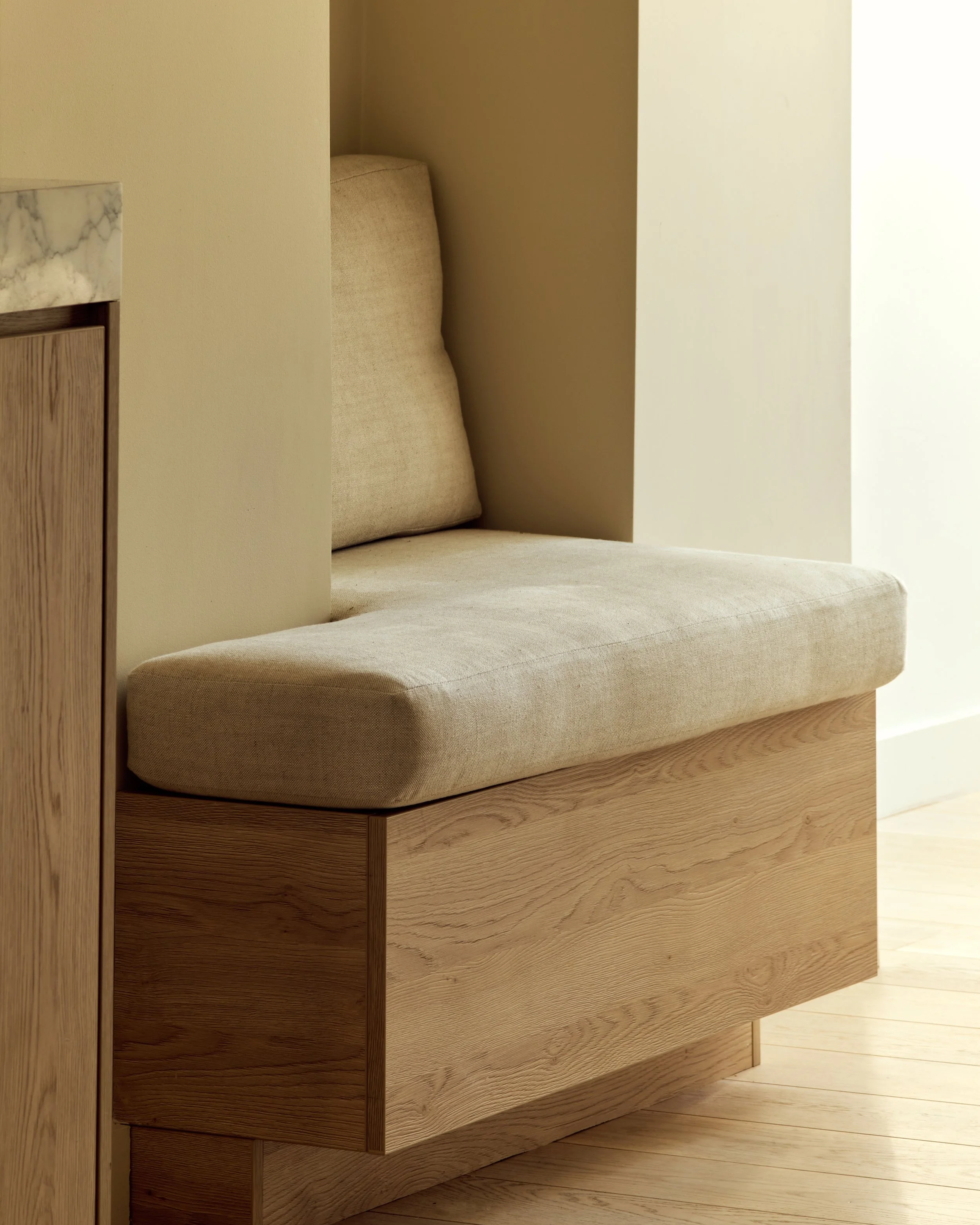 A beige upholstered built-in bench with a cushion, positioned under a small nook beside a wall and wooden flooring.