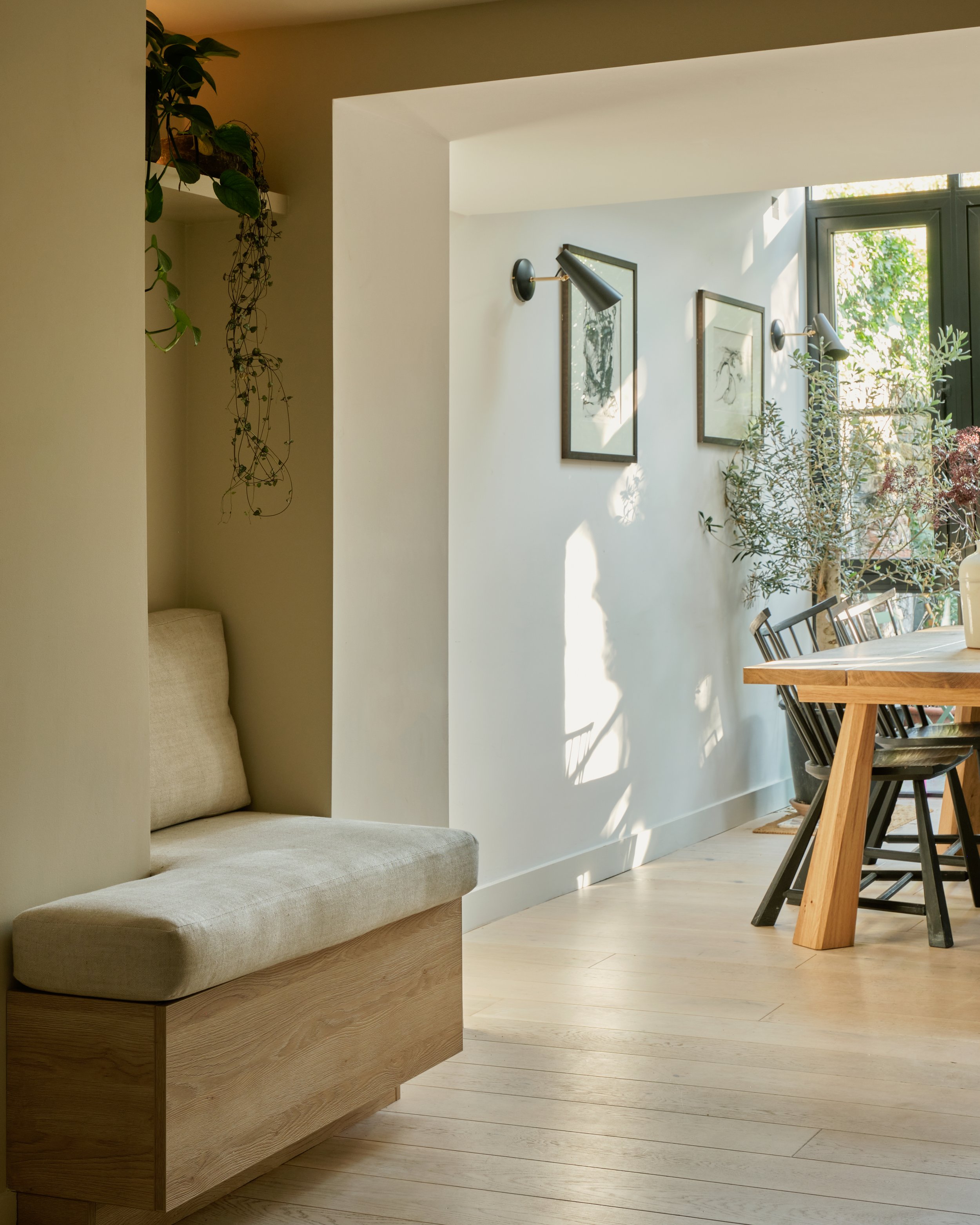 Sunlit dining area with wooden table, black chairs, potted plants, framed artwork, wall-mounted lights, and large window with greenery outside.