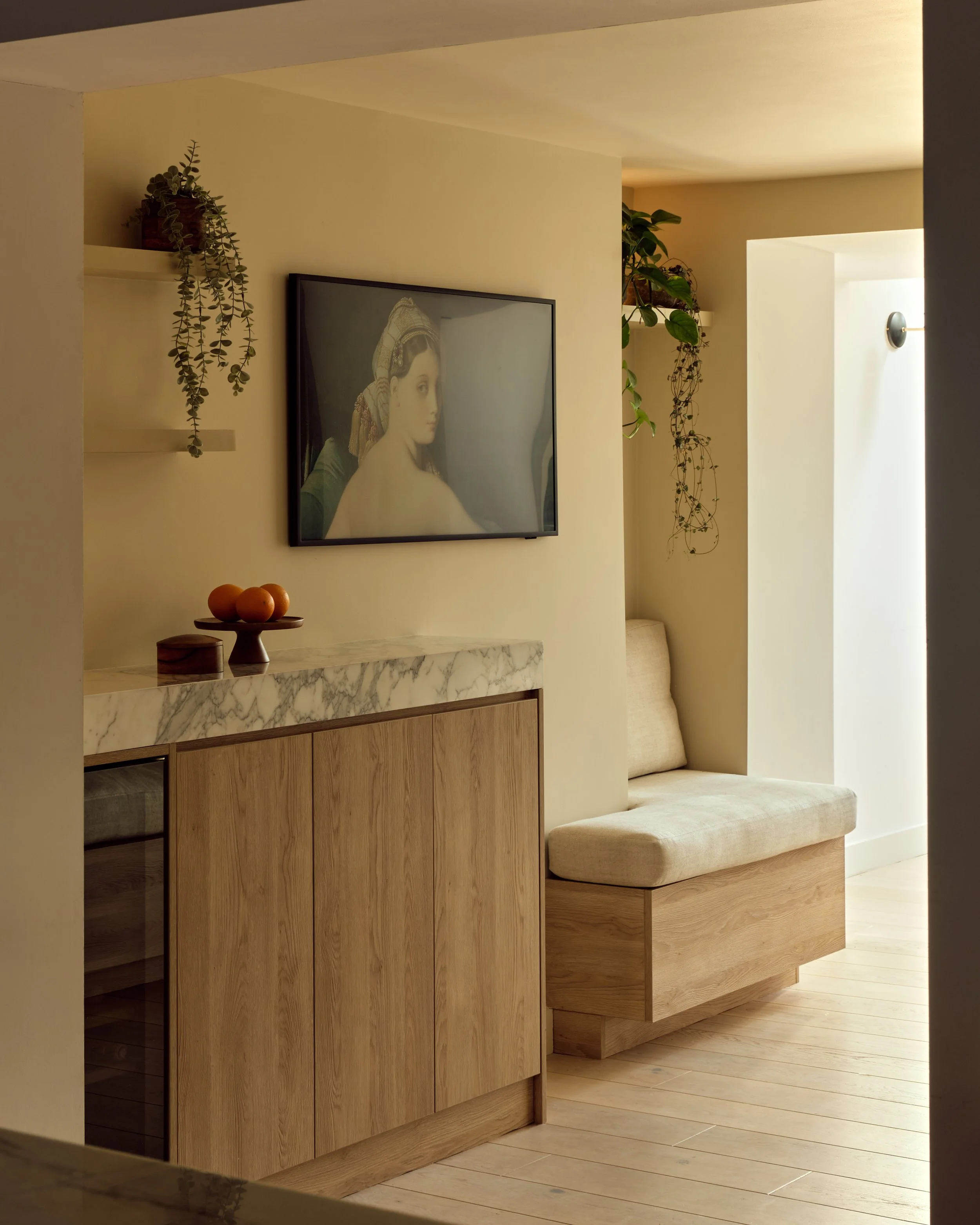 A cozy interior corner of a room with a mounted TV showing a classical portrait, a marble-topped wooden cabinet with oranges on a mini cake stand, a built-in cushioned wooden bench, and hanging plants in a well-lit space with natural light.