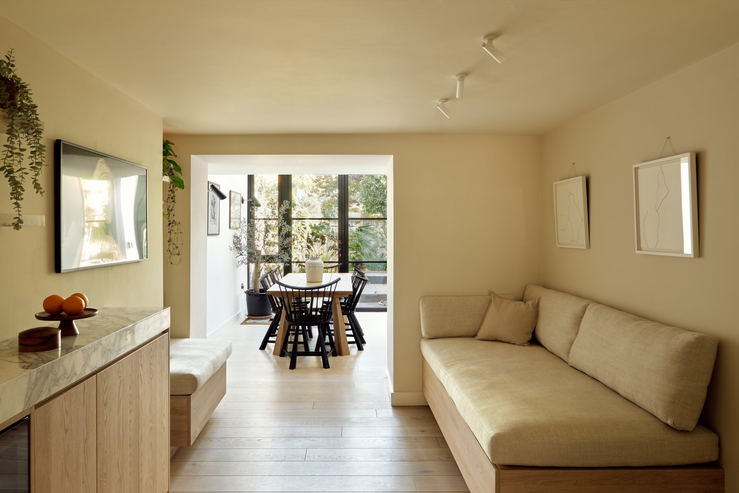 Bright living space with a beige sofa, a marble-topped wooden cabinet with decorative objects, wall-mounted TV, and framed artwork, leading to a dining area with a wooden table and black chairs near large window with plants outside.