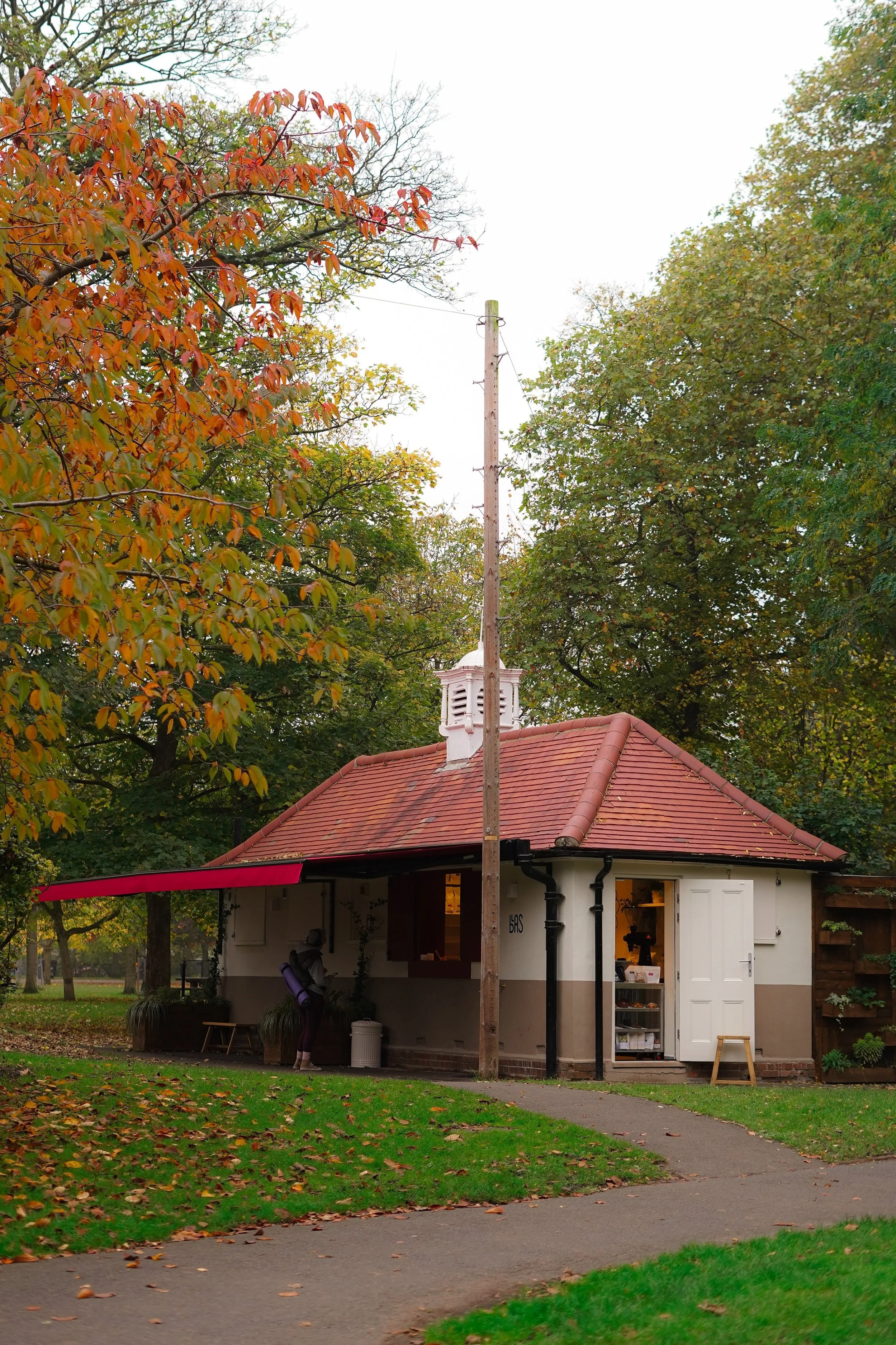 Small white building with a red-tiled roof, surrounded by trees with autumn foliage. One person near the building leans against it, and a sidewalk curves in front of it.