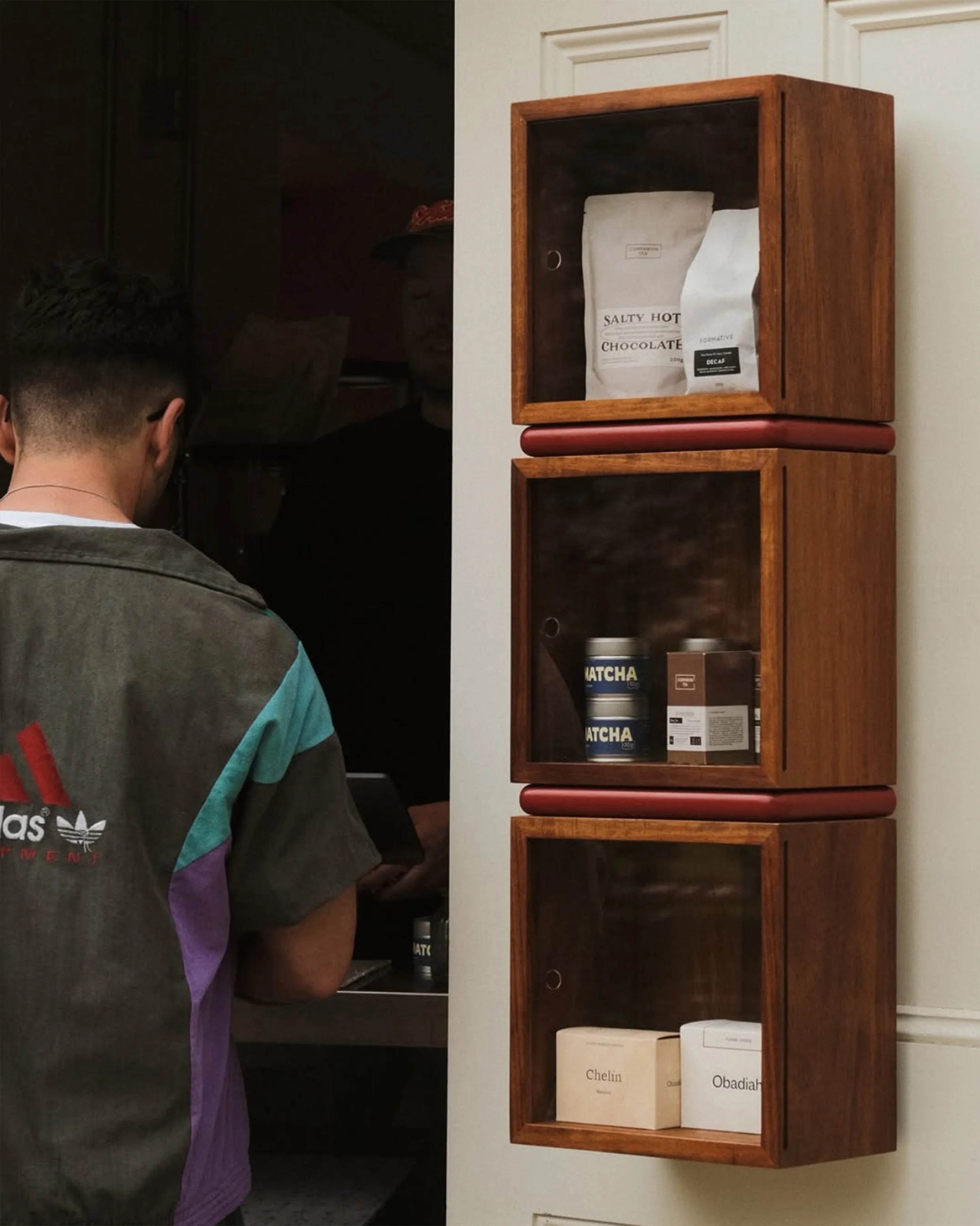 Wall-mounted wooden cabinets with glass doors containing a bag of Salty Hot Chocolate, cans of Matcha, and boxes labeled Chelin and Obadiah, with a person in a colorful jacket standing nearby.