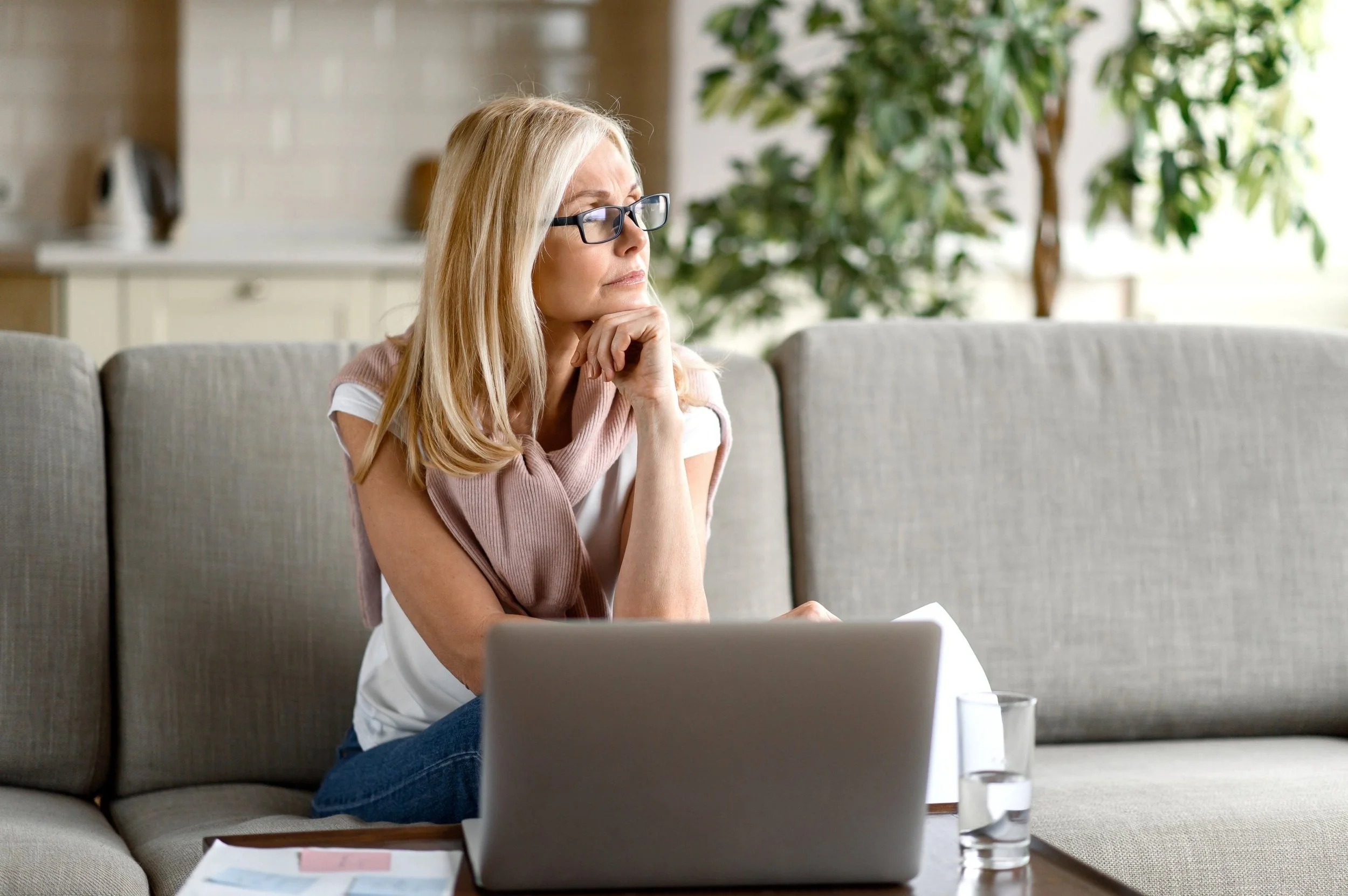 A woman with blonde hair and glasses sitting on a sofa, looking thoughtfully at a laptop in front of her. There is a glass of water and some papers on the table.