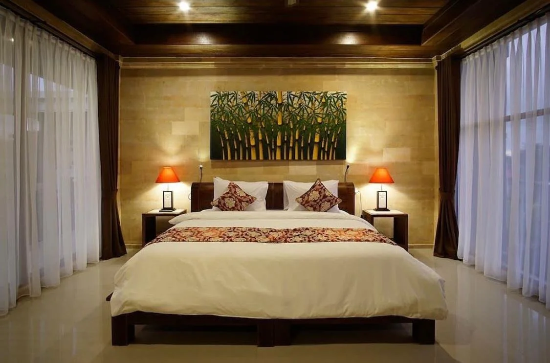Chambre d'hôtel avec un lit double, deux tables de nuit avec lampes à abat-jour rouges, tableau de feuillage au-dessus du lit, rideaux blancs et rideaux bruns aux fenêtres