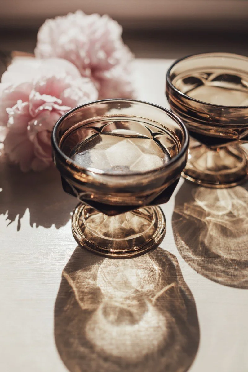 Two glass bowls filled with a dark liquid, placed on a light surface, with pink flowers in the background. Sunlight creates shadows and reflections on the surface.