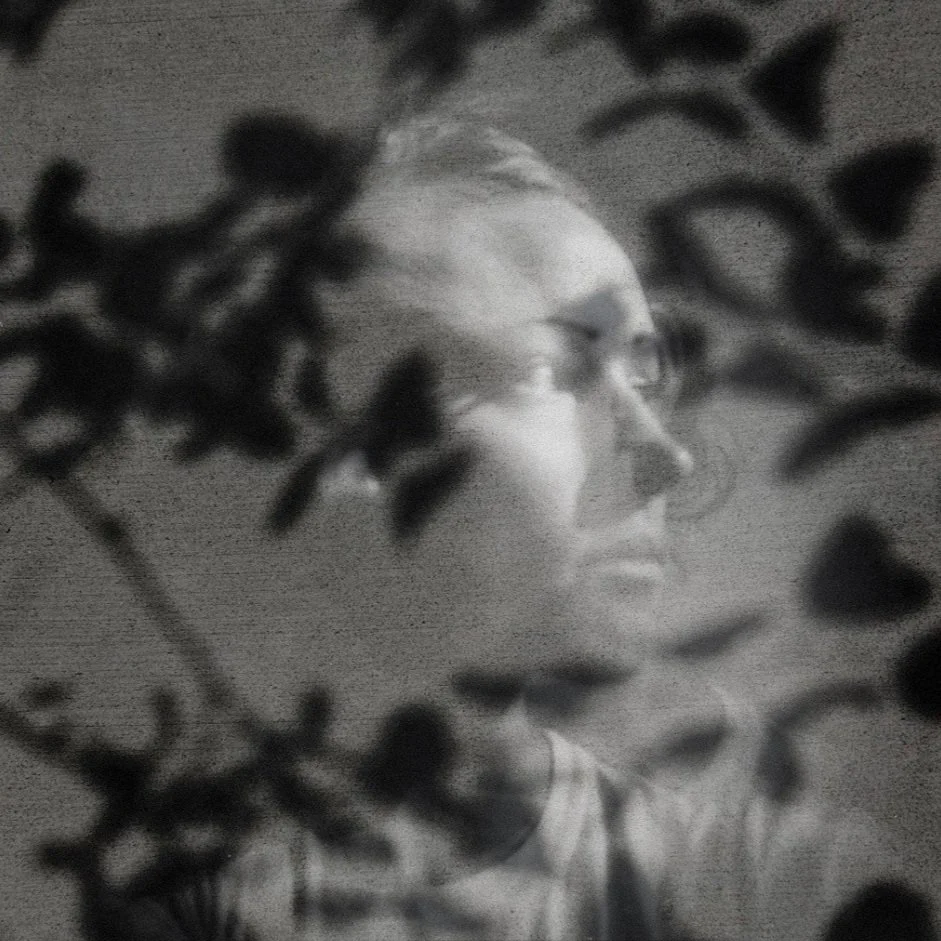 Black and white photo of a woman, seen through leaves in the foreground, looking to the side with glasses and a thoughtful expression.