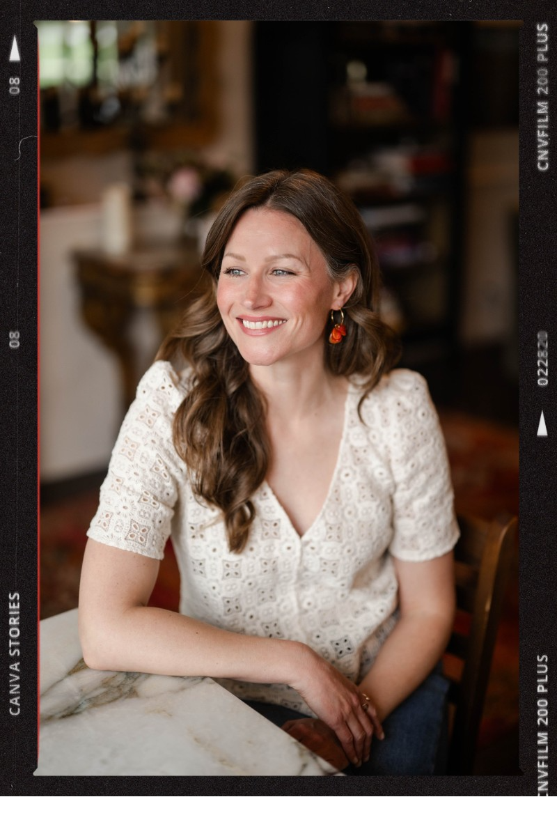 A woman with long brown hair, wearing a white lace blouse and orange earrings, smiling while sitting at a table in a cozy room.