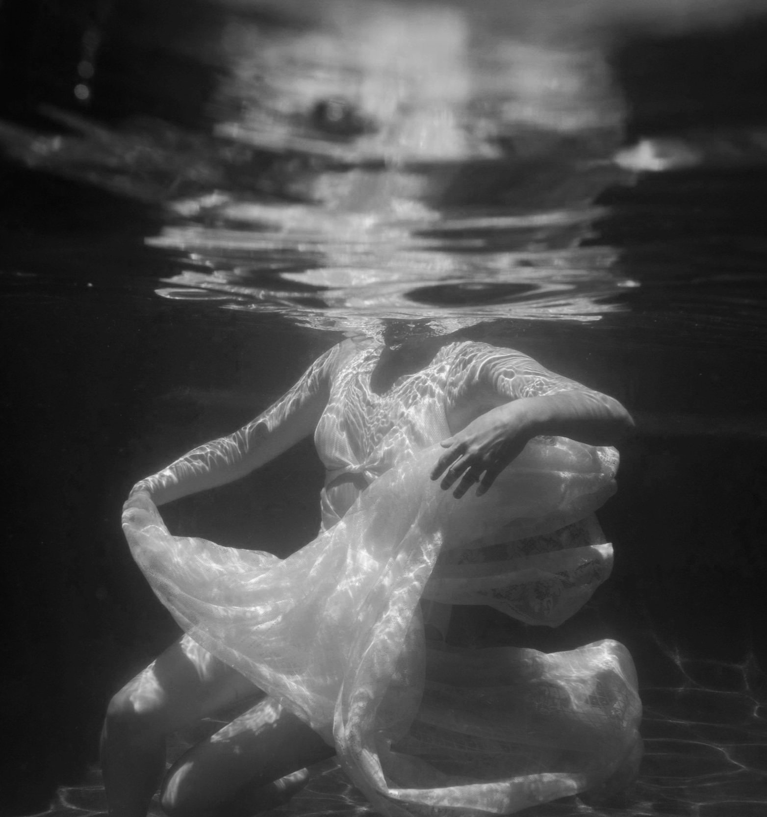Black and white underwater photograph of a woman in a flowing dress, her head submerged, with light reflections on the water's surface.