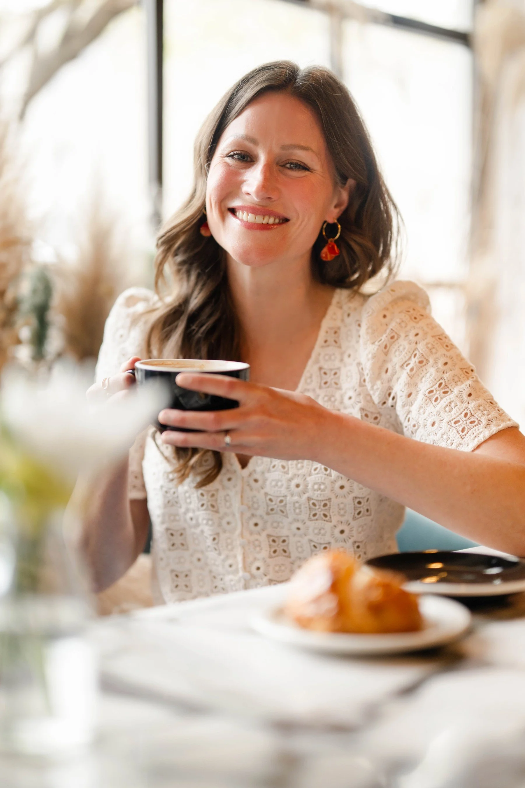 A woman with long brown hair wearing a white eyelet dress, smiling, holding a black mug at a dining table.