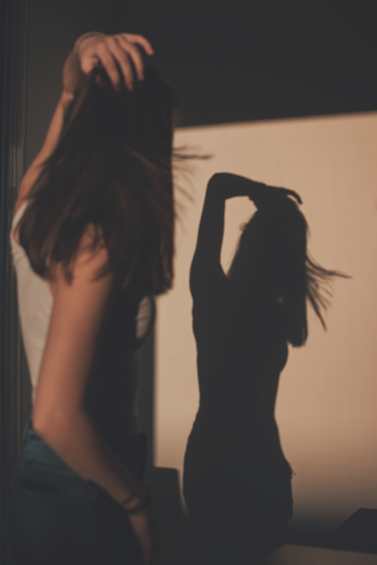 A woman with brown hair, wearing a white top, stands near a wall with her hand in her hair, casting a shadow on the wall that resembles her pose.