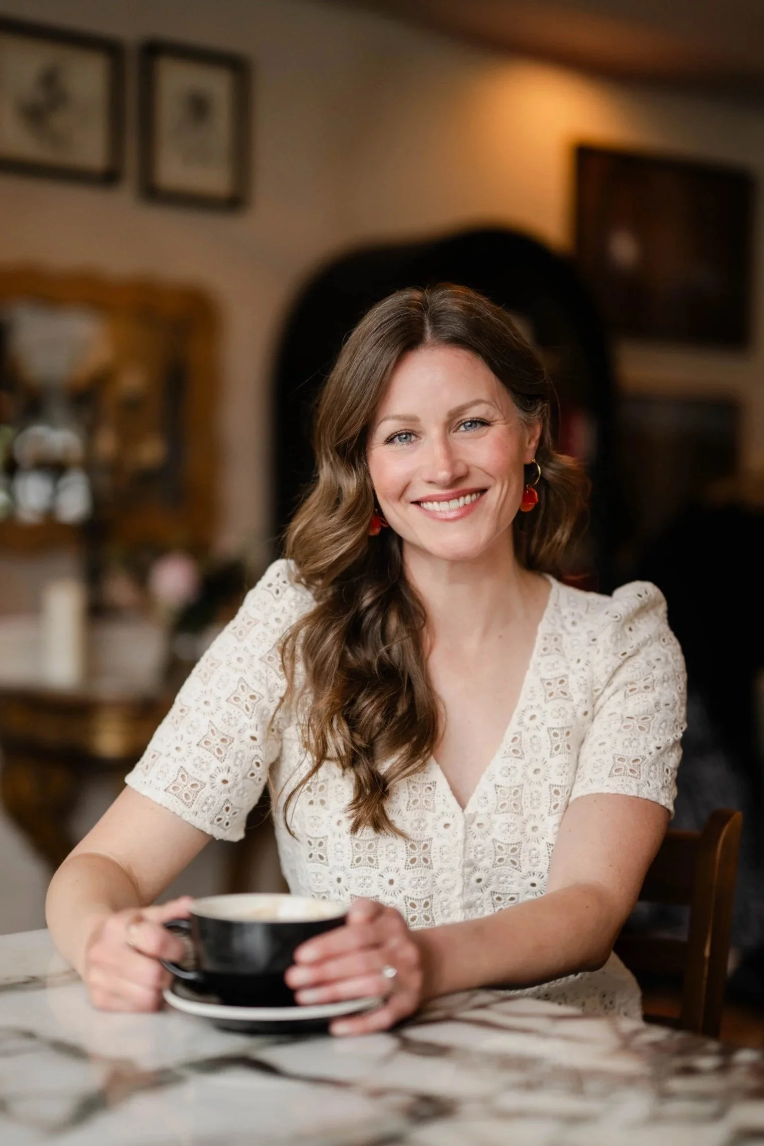 A smiling woman with brown wavy hair, wearing a cream lace top and red earrings, sitting at a table holding a black cup in a cozy café or restaurant.