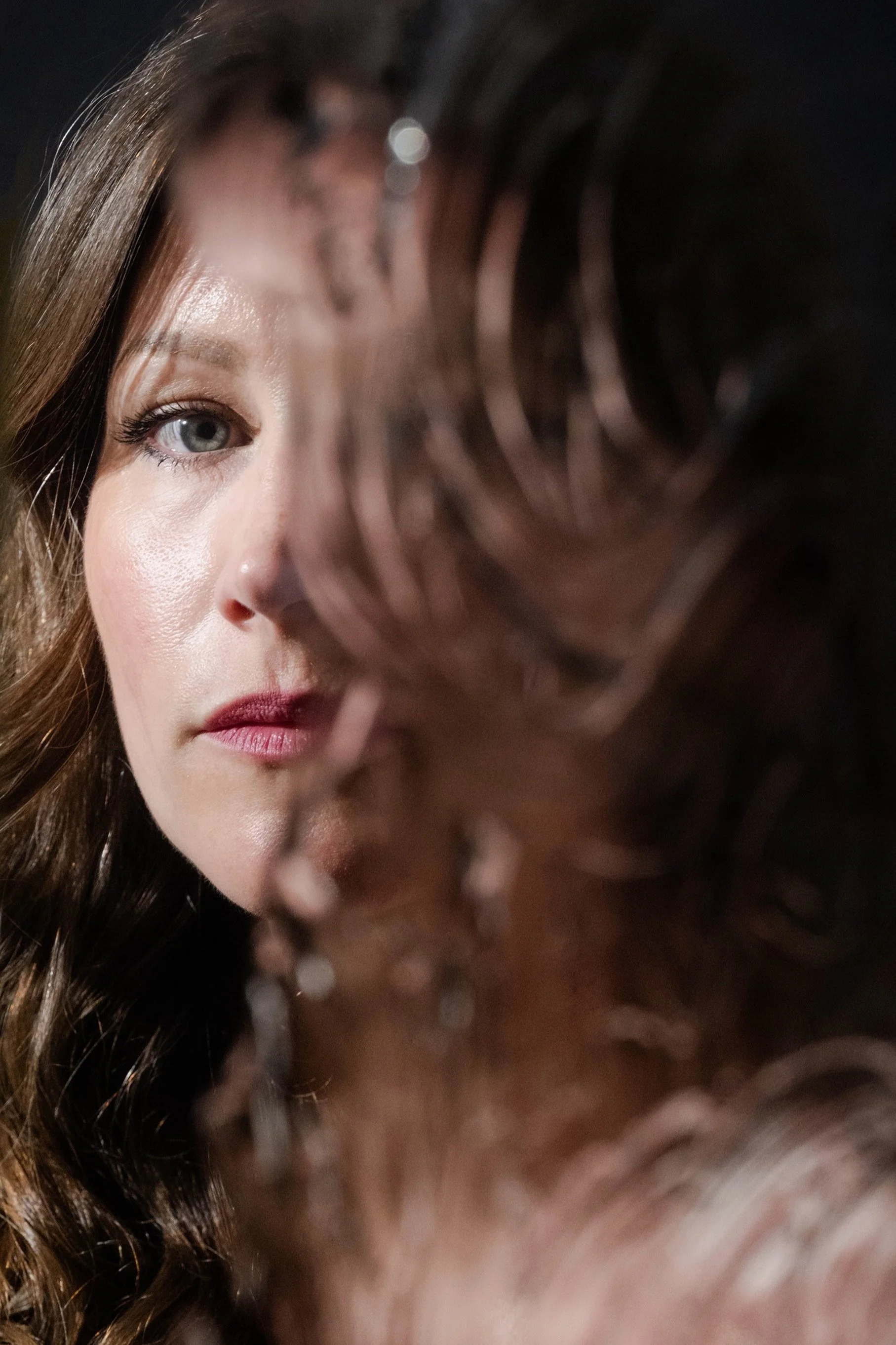 Close-up of a woman's face with blue eyes and brown hair, partially obscured by a blurred, curly hair in the foreground.