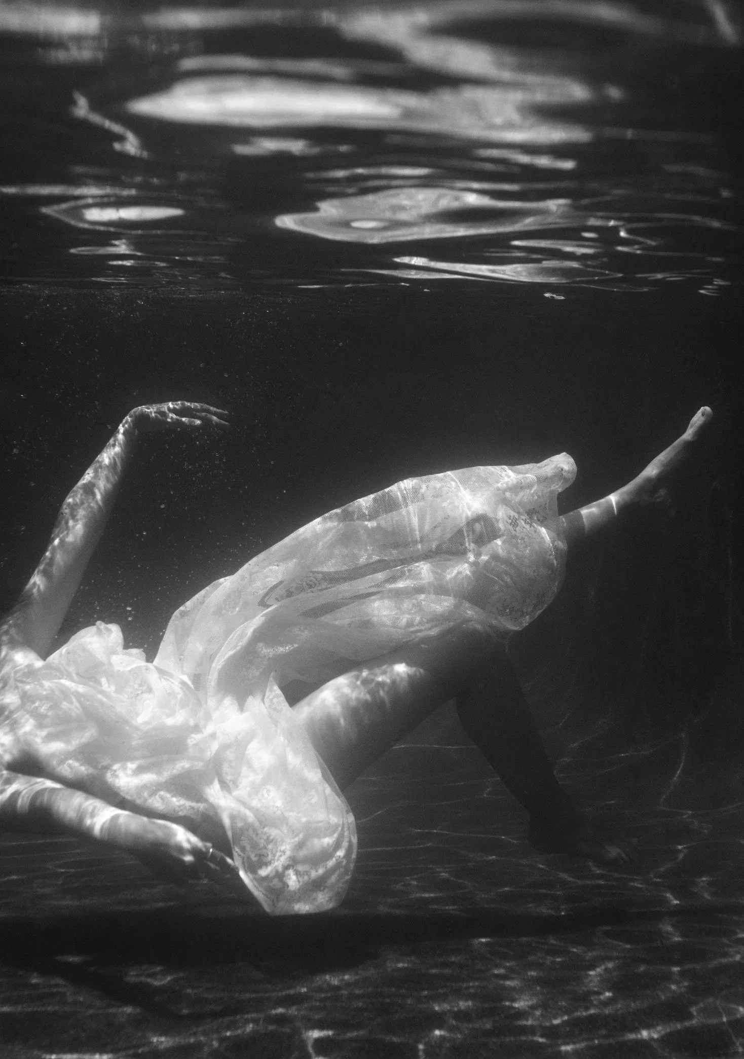 A person underwater, wearing a flowing dress, with their arms extended and head tilted back, captured in black and white.