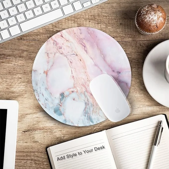 A marble-patterned mouse pad with a wireless mouse on it, on a wooden desk next to a cup of coffee, a cupcake, a keyboard, a tablet, and an open notebook with a pen.