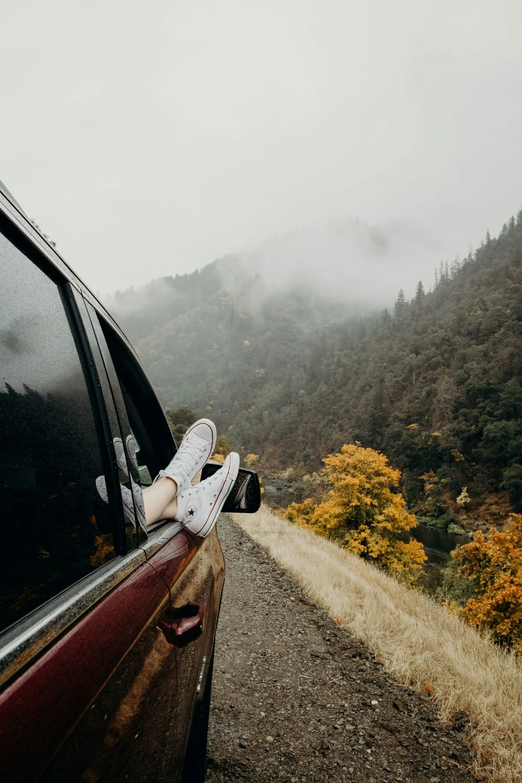 Person with legs outside the car window enjoying scenic mountain view with autumn trees and foggy sky