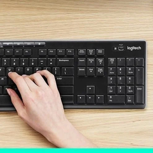Person's hand resting on a black Logitech keyboard on a light wooden surface.