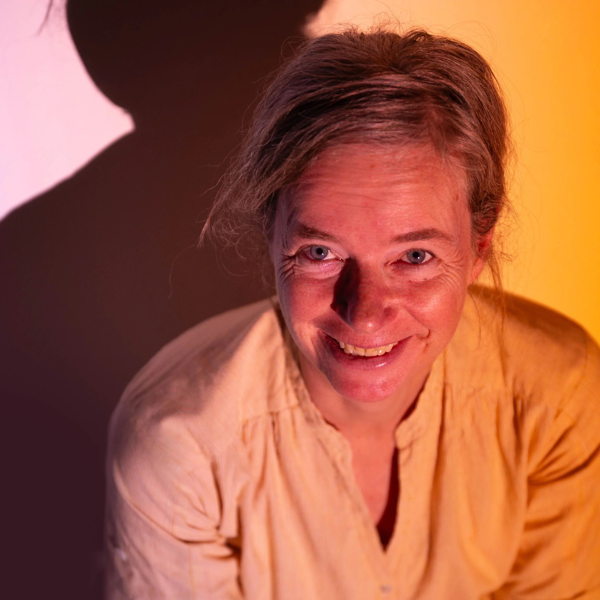 Een lachende vrouw met grijs haar en een beige blouse, gefotografeerd met kleurrijk licht en een oranje en zwarte achtergrond.