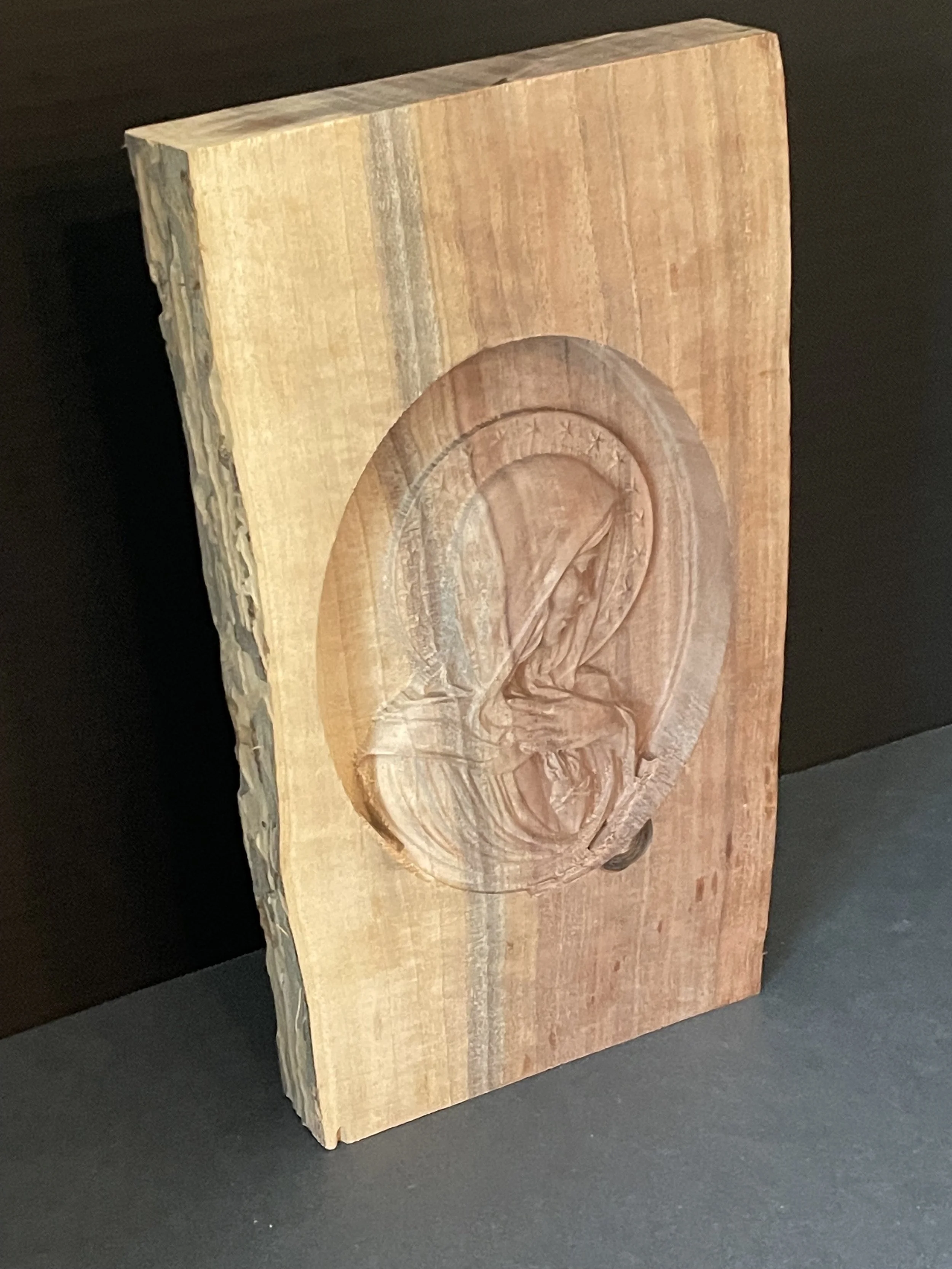 A wooden carving of the Virgin Mary with a halo, holding a baby Jesus inside an oval frame, all carved into a single piece of wood.