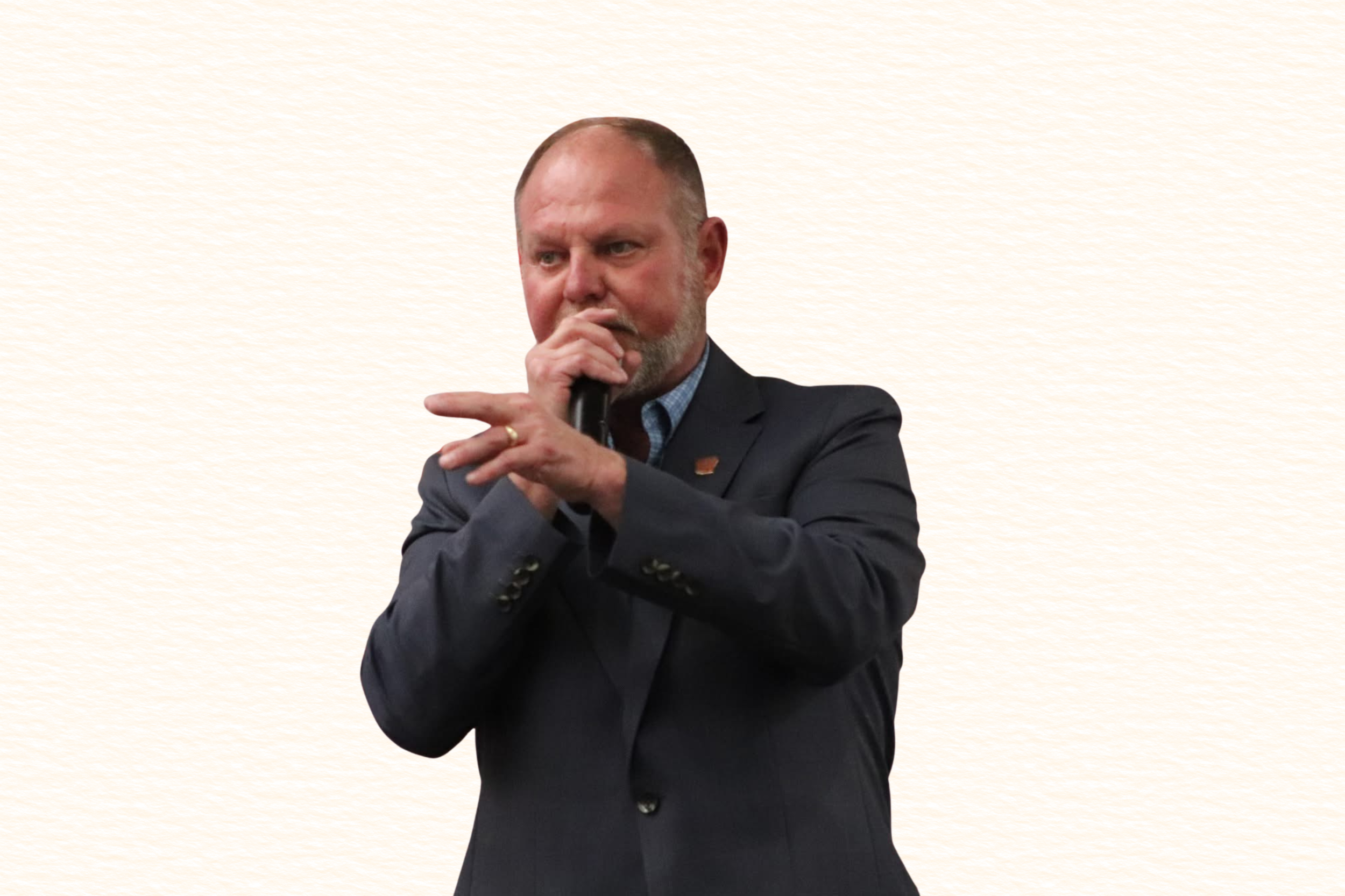 A man in a dark suit holding a microphone near his mouth, standing against a light-colored background.