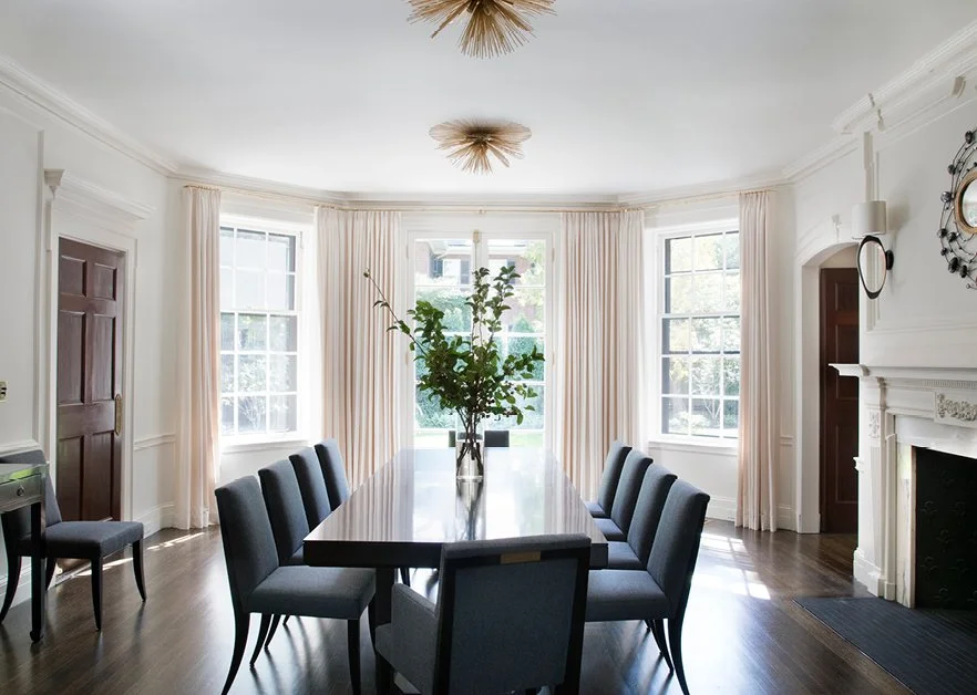 Bright dining room with large windows, a dark wood table, ten grey upholstered chairs, a tall potted plant centerpiece, white curtains, a dark wooden door, a white fireplace, and decorative wall accents.