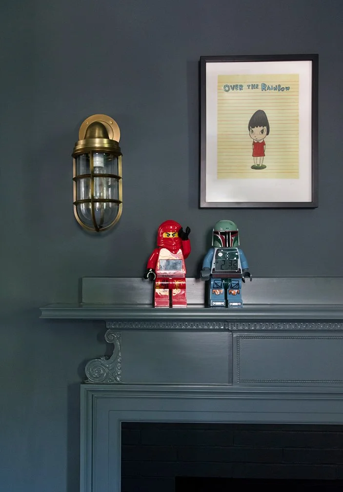 Two LEGO Star Wars figurines on a gray mantle, one dressed as a female character in red and the other as Boba Fett, with a framed drawing of a girl titled "Over the Rainbow" on a dark gray wall and a brass wall sconce to the left.