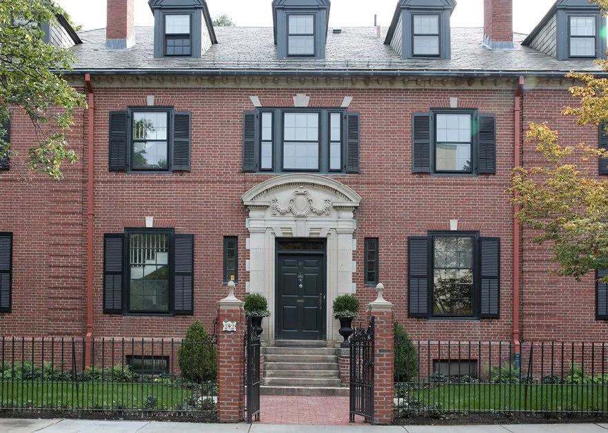 Front view of a red brick house with black-shuttered windows, a dark green front door, and a gated entrance with steps leading to the door, surrounded by a small garden and trees.