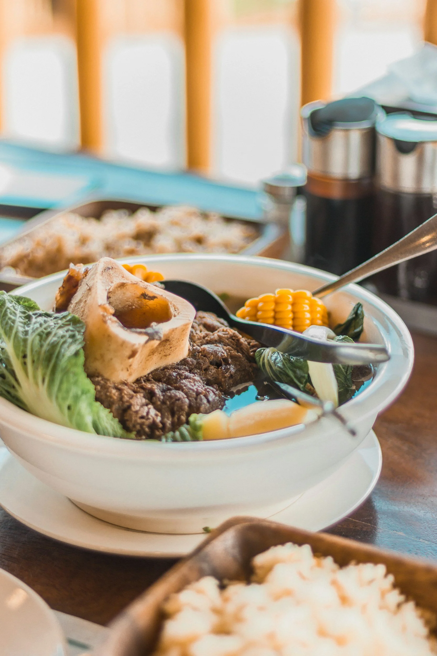 A bowl of Asian beef noodle soup with vegetables, corn, and a soft-boiled egg on top, placed on a wooden table with rice and condiments in the background.