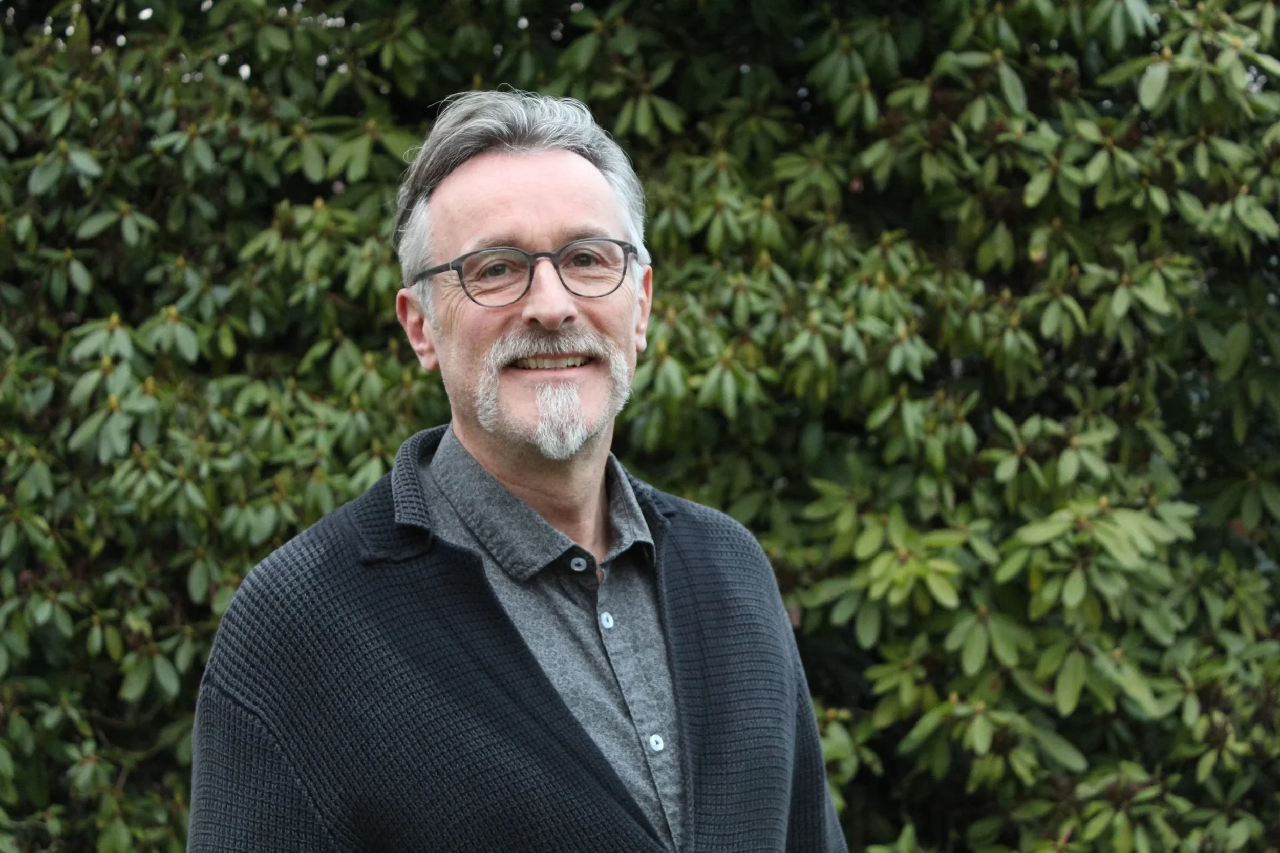 Ein lächelnder Mann mit Brille, grauem Haar und Bart steht vor einer grünen Laubwand im Freien.