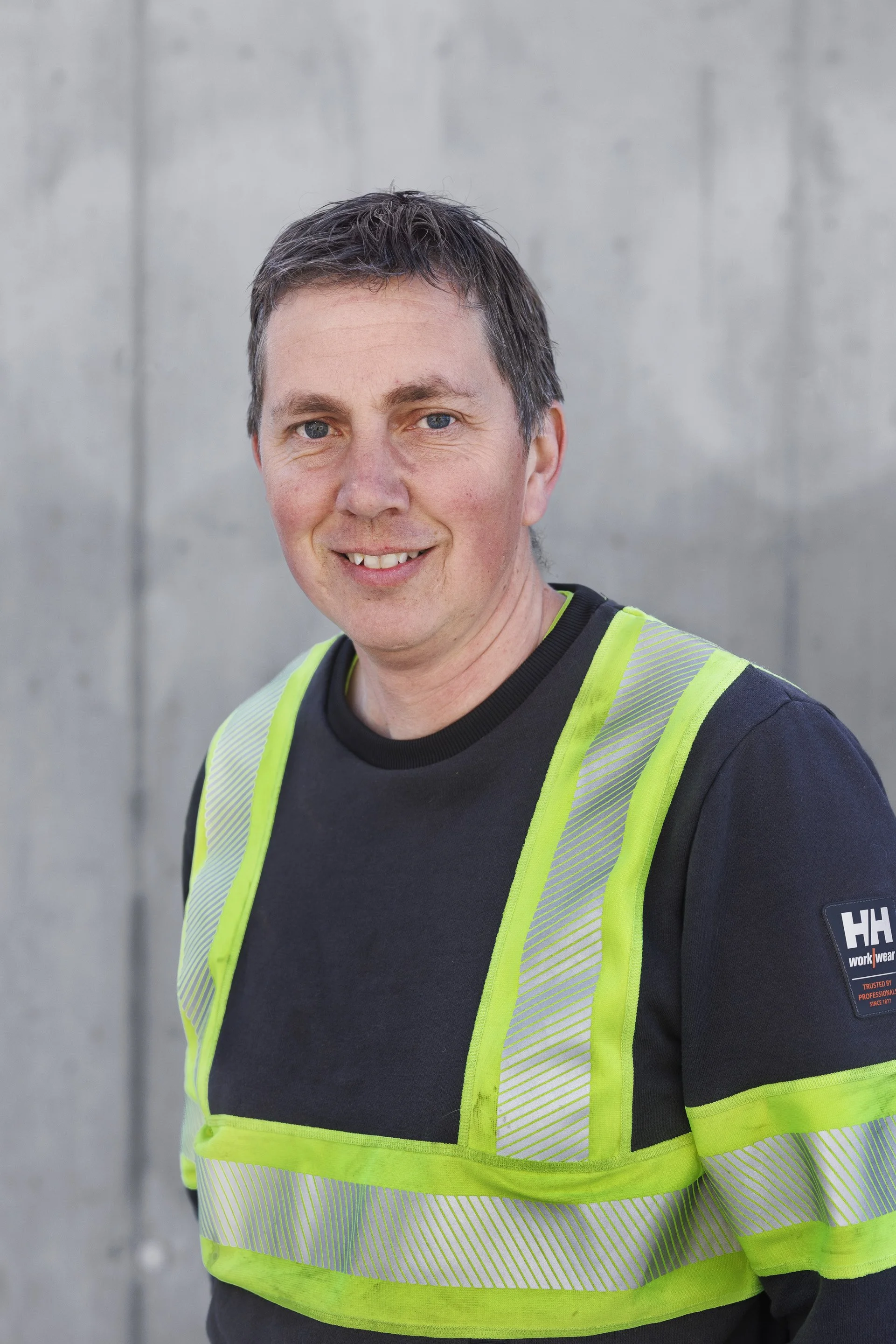 Mann iført svart t-skjorte og høyrefeltsvest står foran en betongvegg.