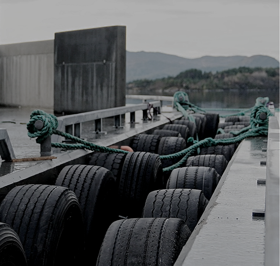 Bilde av en flytekai med fendere og tjukke tau som er festet til fortøyningsboller, med vann og fjell i bakgrunnen.