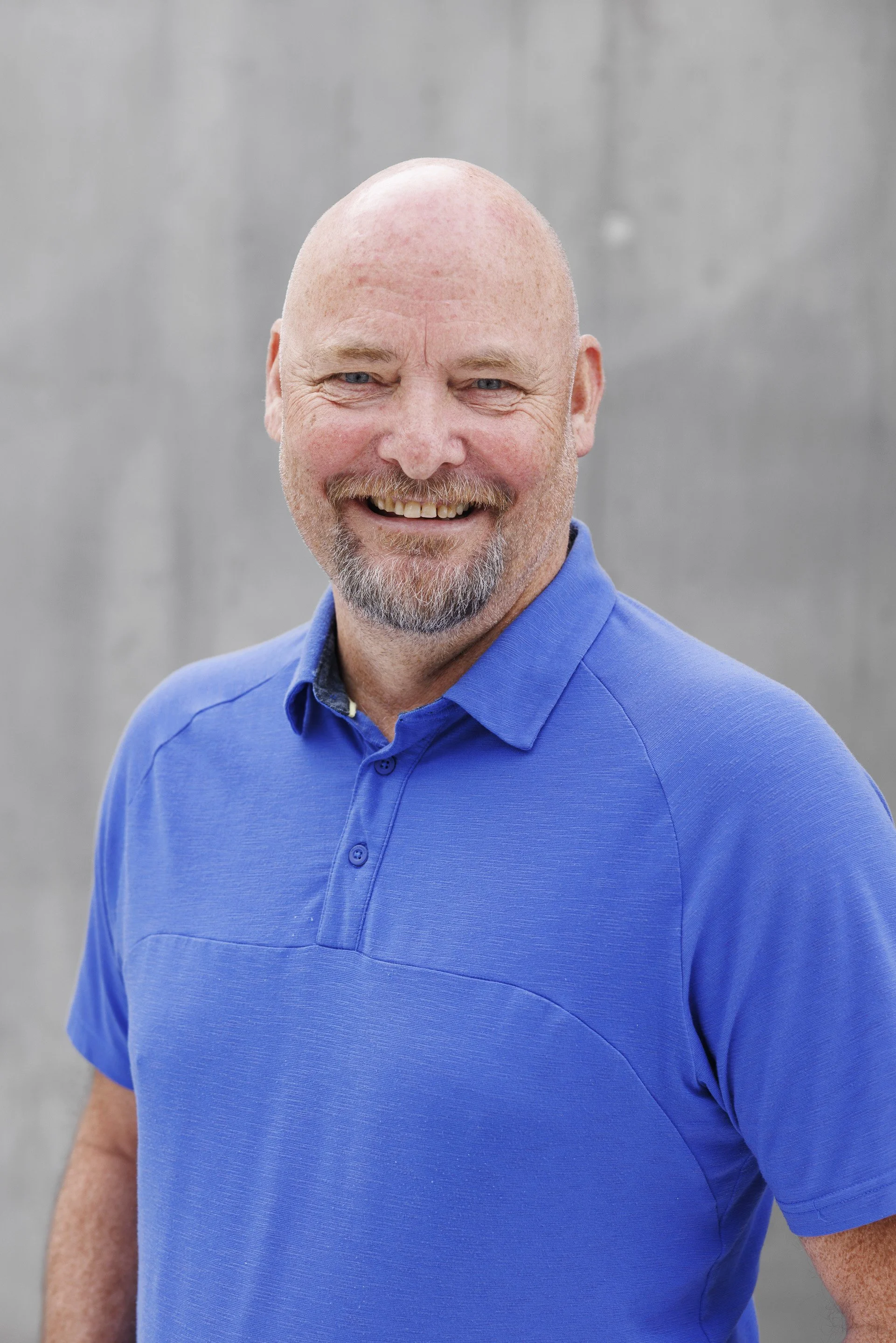 Portrett av en smilende eldre mann med skjegg, iført blå polo shirt, mot en grå betongvegg.