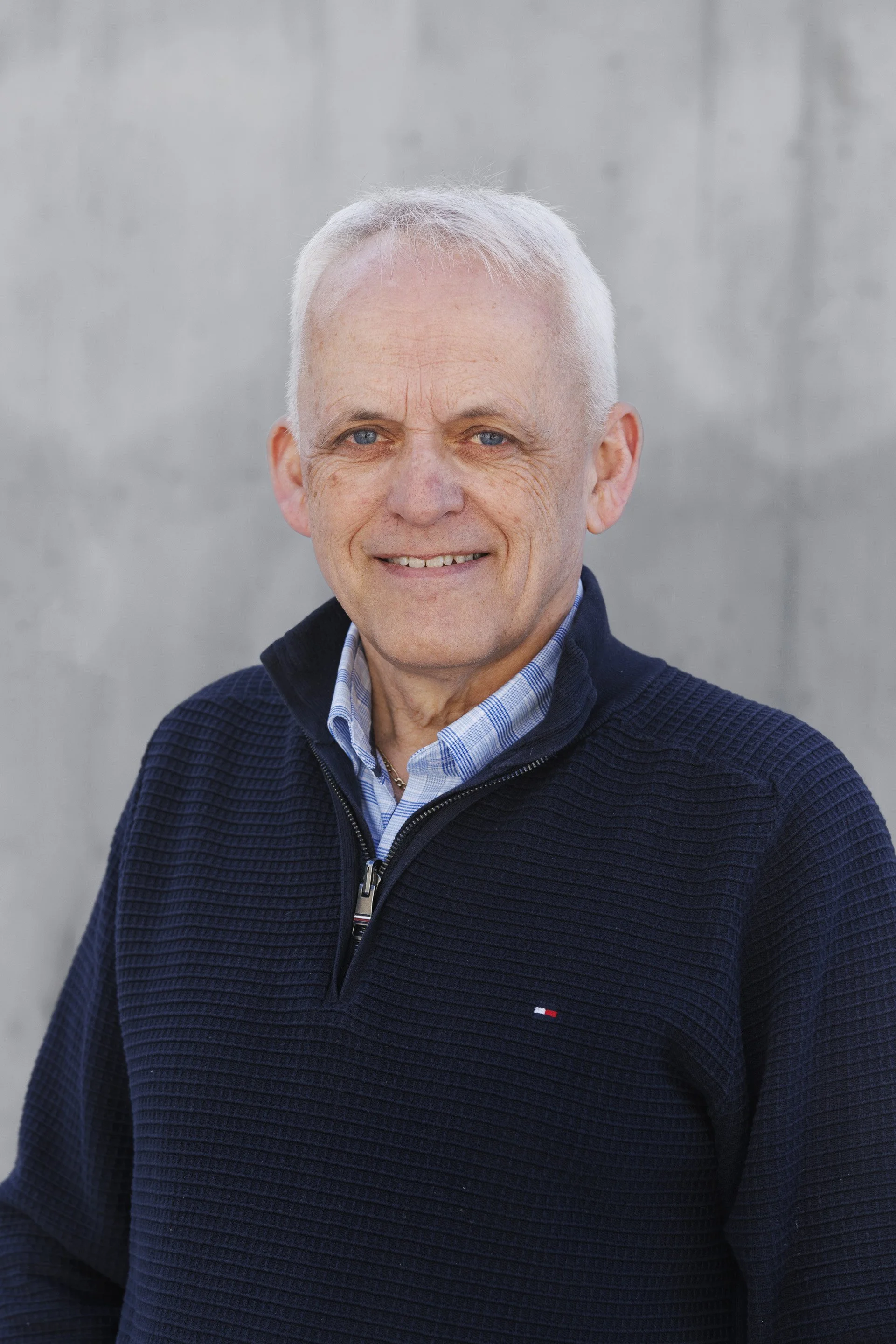 Et portrett av en eldre mann med kort, hvitt hår, som smiler og har på seg en mørk blå genser og en lys blå skjorte.