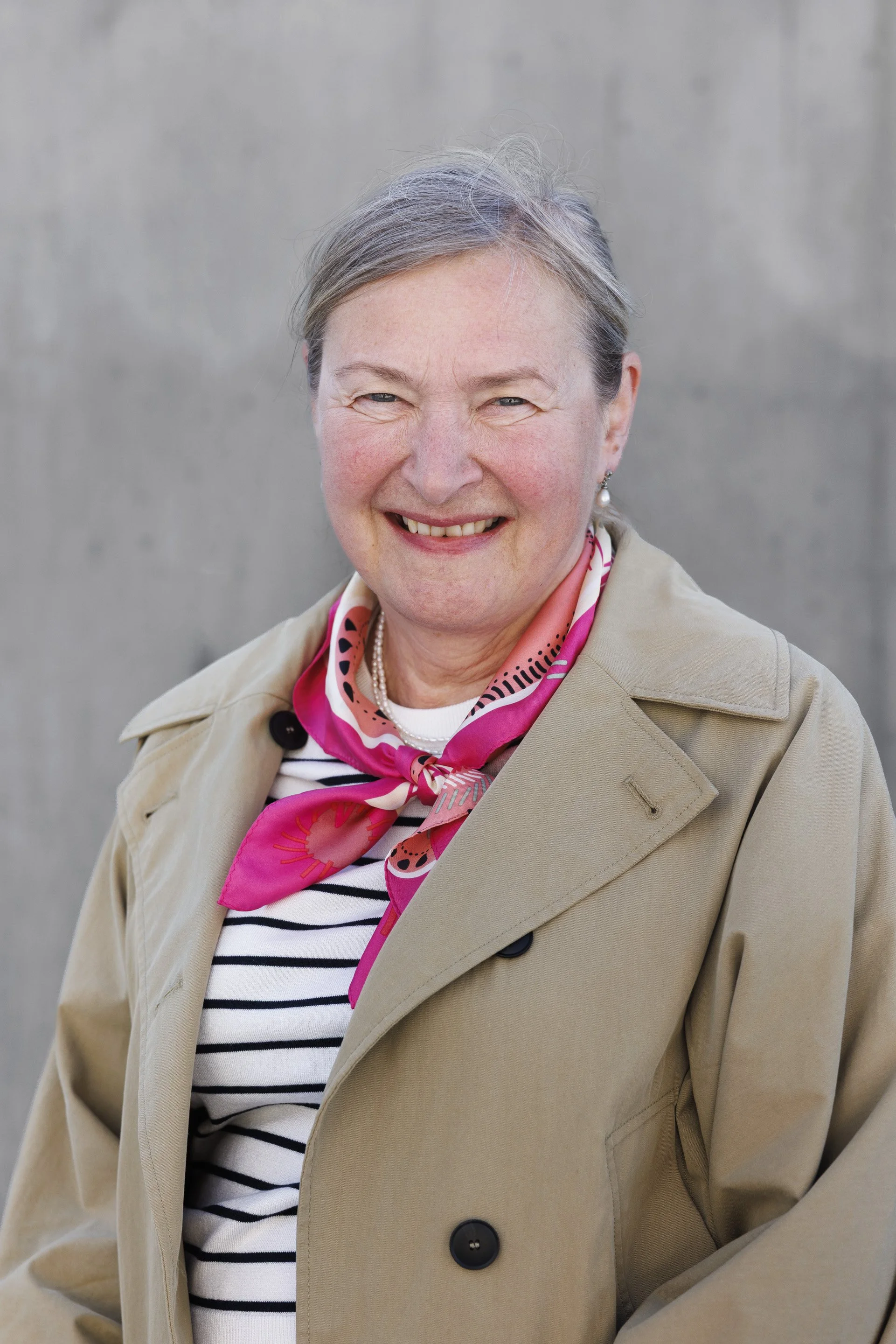 Et portrett av en eldre kvinne med grått hår, smilende, iført beige trenchcoat og rosa og hvit silkeskjerf, mot en grå bakgrunn.