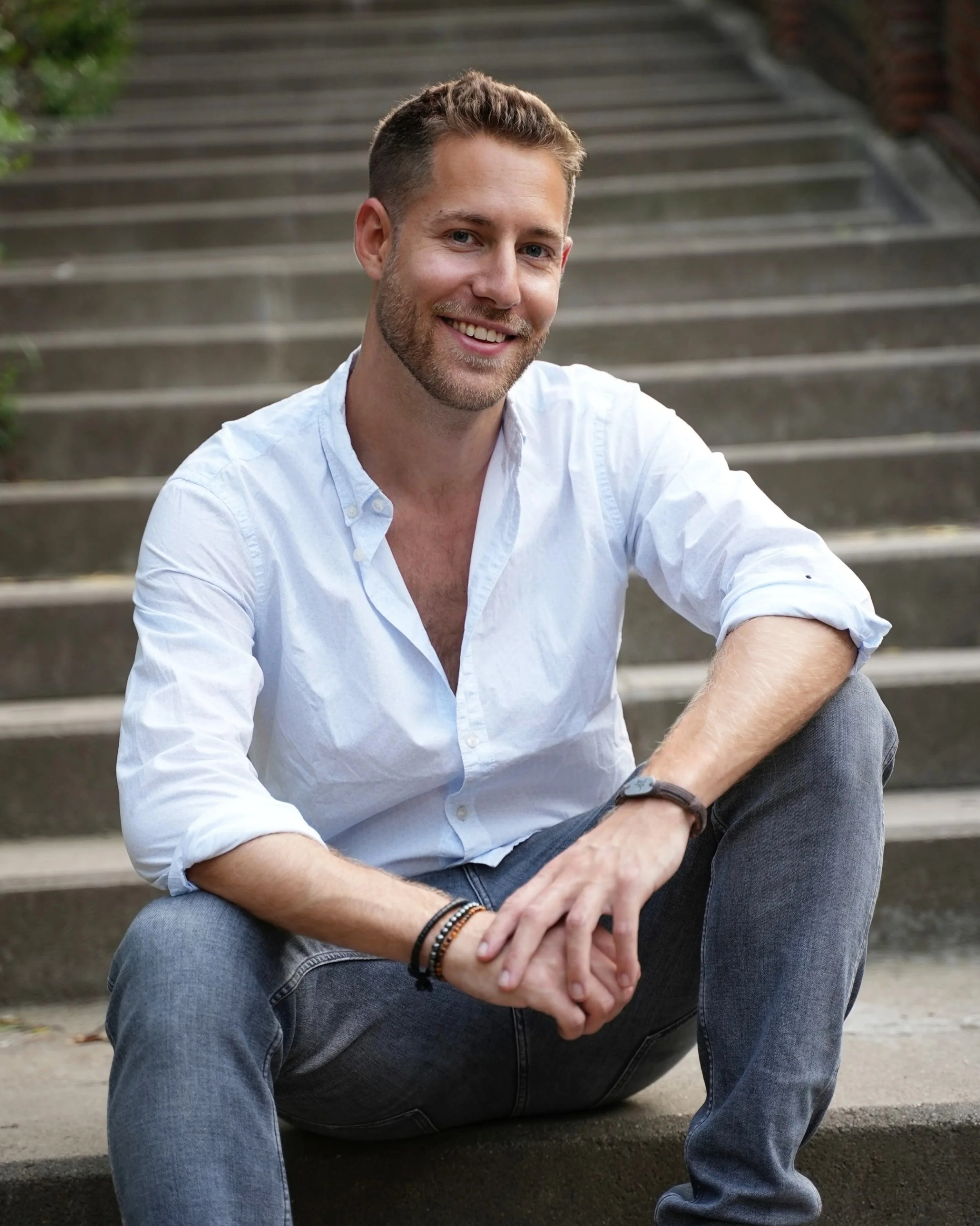 Shai Schechter wearing a white button-up shirt with the sleeves rolled to the elbows, dark jeans, and a wristwatch. He is smiling and looking at the camera.