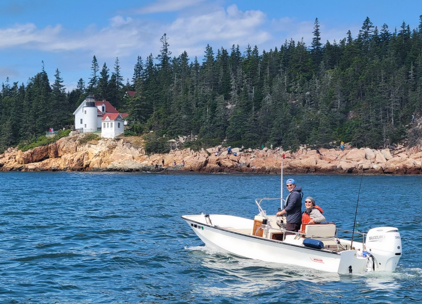Two people on a small white motorboat with a Suzuki engine, cruising on blue water, with a rocky shoreline and dense green trees, and a white lighthouse with a red roof in the background.