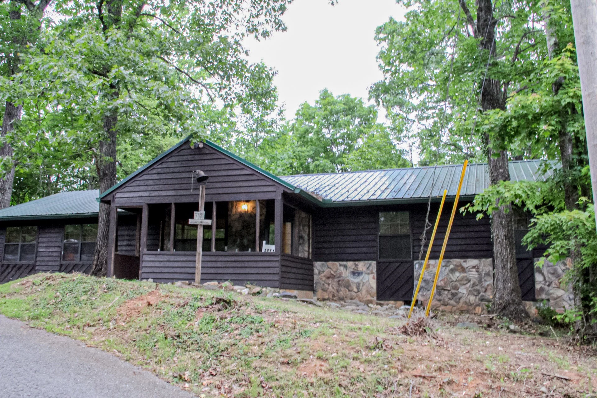 A black wooden house with a stone foundation, metal roof, and a porch with a light on inside, surrounded by green trees.